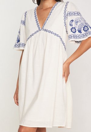 Biała sukienka do kolan z głębokim dekoltem w serek i niebieską kwiatową haftem na rękawach i dekolcie, noszona przez osobę z ręką w kieszeni.