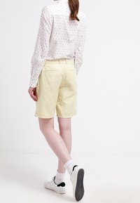 Shorts jaune clair, longueur genou, en tissu doux, avec un devant plat, des poches latérales et une coupe décontractée. Portés avec une chemise à manches longues à motifs et des baskets blanches.