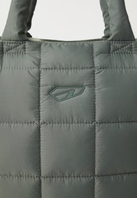 Sac fourre-tout vert rembourré avec texture matelassée, présentant des coutures surpiquées et un accent de logo moulé. Poignées renforcées pour le transport.