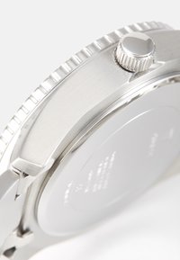 Sudraba nerūsējošā tērauda pulkstenis ar teksturētu rāmīti un skrūvējamu koroni; ir gluda, atstarojoša aizmugure.