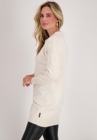 Crème-kleurige trui met lange mouwen, zachte textuur en zijsplitten. Gedragen boven zwarte leren legging, met een klein merkembleem.