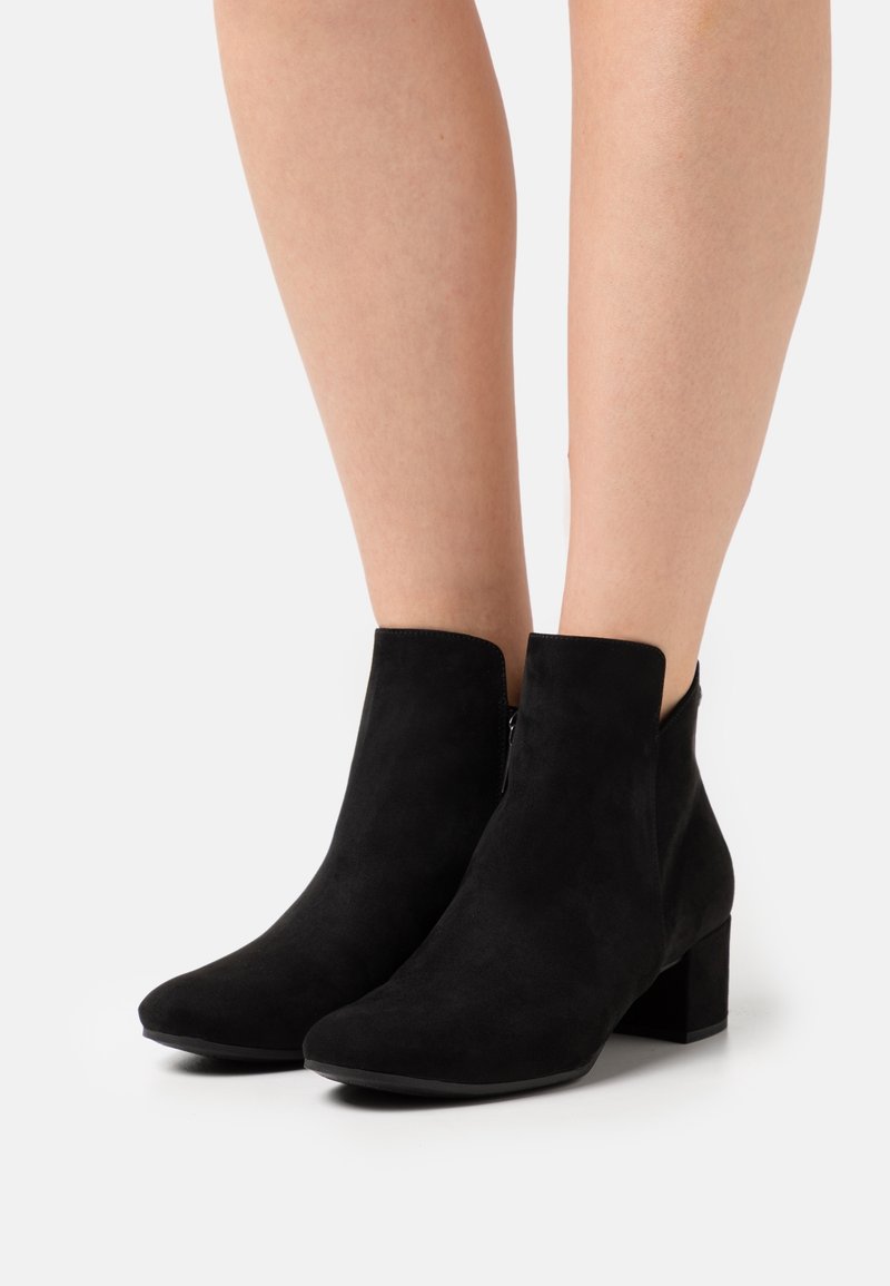 Schwarze Wildleder-Stiefeletten mit Blockabsätzen, die auf nackten Beinen vor einem schlichten weißen Hintergrund getragen werden.