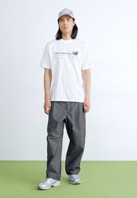 T-shirt blanc à manches courtes avec le logo noir "New Balance Athletics", pantalon gris ample et baskets grises avec des accents réfléchissants.