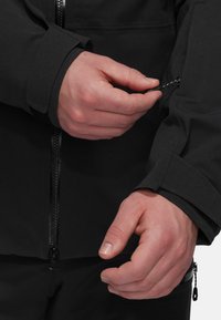 Black soft-shell jacket featuring a zippered cuff and a smooth texture. Hands adjusting the cuff's closure with a discreet zipper detail.