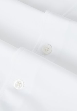 Fehér ingujjközeli kép, varrott gomblyukakkal és fehér műanyag gombokkal, világos, puha megvilágításban.