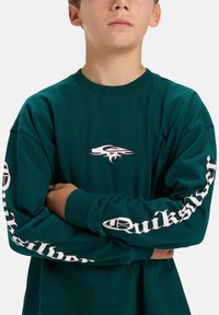 Groene longsleeve t-shirt van katoen met een ronde hals. Kenmerkt zich door een wit logo op de borst en verticale tekst op de mouwen.