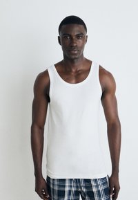 Top sin mangas de algodón blanco con tirantes anchos, de diseño ajustado. El modelo lleva puestos unos pantalones cortos a cuadros en tonos de azul y gris.