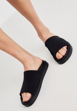 Fødder iført sorte slip-on sandaler med tykke såler på en lysegrå baggrund.