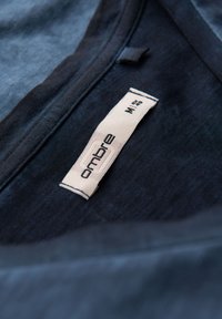 Tmavě modré tričko s kulatým výstřihem, textura látky je hladká. Viditelná nálepka s nápisem "ombre", velikost M, s šitím podél výstřihu.