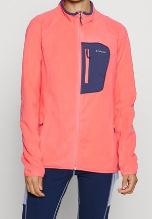 Bright coral zip-up fleece jacket with navy chest pocket and Columbia logo, worn with navy pants featuring light gray side panels.