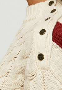 Cremefarbener Kabelstrickpullover mit geripptem Kragen und bronzefarbenen Knöpfen, der ein weinrotes Panel aufweist. Das strukturierte Muster ist auf dem Stoff sichtbar.