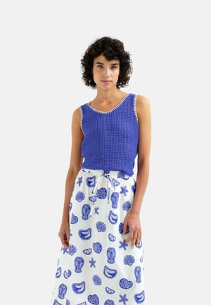 Mujer con el pelo corto y rizado que lleva un top sin mangas de punto azul y una falda blanca con estampados azules de criaturas marinas y conchas.