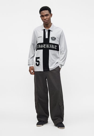 Jeune homme portant un maillot de rugby blanc avec une grande croix noire et un pantalon cargo noir, debout devant un fond blanc uni.