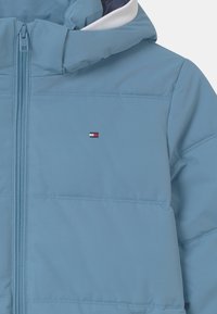 Světle modrá zateplená bunda s předním zipem, která má malou vyšívanou nášivku a kontrastní podšívku v tmavší barvě.
