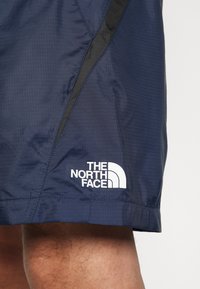 Shorts bleu marine en tissu léger, avec un panneau latéral noir et un logo blanc de The North Face sur l'ourlet inférieur.