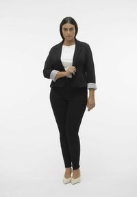Blazer noir avec des poignets rayés retroussés, porté sur un top blanc et associé à un pantalon noir ajusté. Le modèle porte des escarpins beiges à bout pointu.