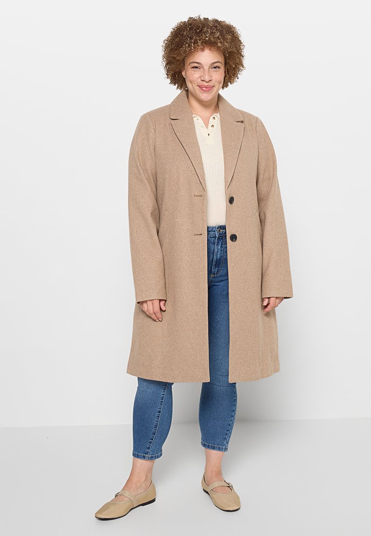 Beige Wollmischung langer Mantel mit strukturiertem Revers, doppelt-knopfverschluss und seitlichen Taschen, getragen über einem cremefarbenen Oberteil und blauen Jeans.