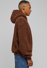 Ruda vilnos gobtuvė su tekstūruota paviršiumi, gobtuvu ir elastingais rankogaliais, derinama su mėlynais džinsais. Šoninis profilio vaizdas.