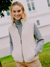 Femme souriante aux cheveux blonds courts, portant un gilet en polaire beige sur un pull à col roulé gris, debout en plein air les mains dans les poches.