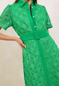 Abito verde in pizzo con maniche corte, caratterizzato da una texture a fantasia, fronte con bottoni e cintura coordinata in vita.