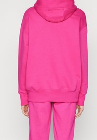 Felpa in pile rosa con un ampio cappuccio, polsini e orlo a coste. Il materiale sembra morbido e testurizzato, con un design semplice senza fantasie.