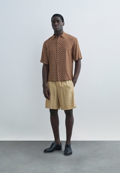 Un homme se tient contre un mur gris, portant une chemise à manches courtes marron à motifs, un short beige à motifs et des chaussures habillées noires.