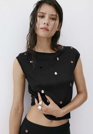 Mujer joven con el pelo mojado lleva una camiseta negra sin mangas con pequeños adornos redondos y brillantes y grandes anillos plateados en los dedos.