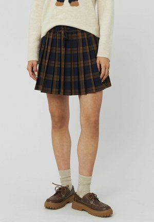 Jupe à carreaux dans des tons marine, marron et crème ; design plissé ; taille avec cordon de serrage ; associée à des chaussures marron et des chaussettes beiges.