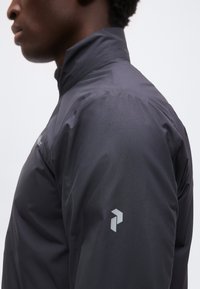 Άτομο που φοράει σκουρόγρια μπουφάν με ψηλό γιακά και μικρό, ανοιχτό γκρι γεωμετρικό λογότυπο στον επάνω βραχίονα, κοιτάζοντας αριστερά σε απλό φόντο.