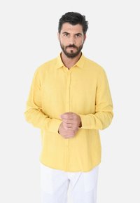 Camicia gialla con bottoni, realizzata in tessuto leggero; presenta un colletto classico, maniche lunghe e una vestibilità comoda. Indossata con pantaloni bianchi.
