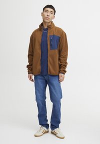 Brun fleecejakke med en blå brystlomme, paret med en blå t-skjorte og blå jeans. Hvite sko med brune detaljer.