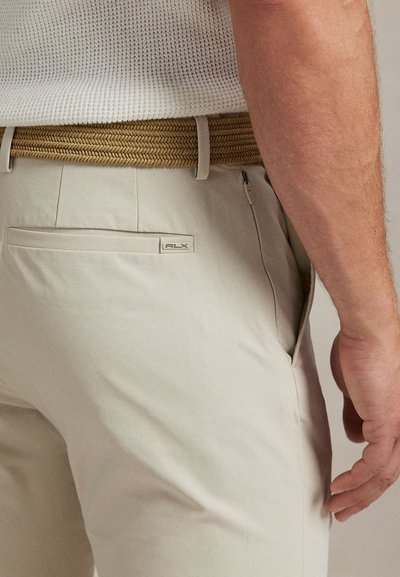 Μπεζ παντελόνι από απαλό ύφασμα, με πίσω τσέπη που φέρει ετικέτα με λογότυπο και πλεκτή ταμπά ζώνη, μερικώς ορατή. Η υφή φαίνεται απαλή.