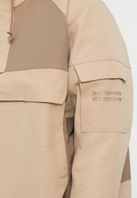 Vêtements d'extérieur beiges présentant un col haut, un design bicolore avec un tissu lisse et des accents texturés, ainsi qu'un logo brodé sur la manche.
