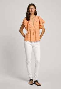 Blusa color pesca a maniche corte con bordi arricciati, motivo ad occhiello, scollatura a V e bottoni decorativi. Abbinata a jeans skinny bianchi e sandali.