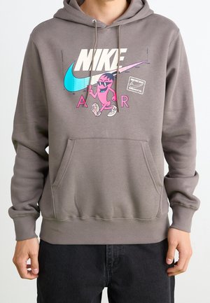 Person iført grå Nike hættetrøje med farverig grafik af en lyserød figur, blå swoosh og teksten "NIKE AIR" foran.
