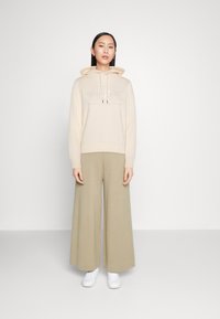 Felpa con cappuccio crema con logo in rilievo, abbinata a pantaloni larghi beige lunghi. Il modello indossa scarpe da ginnastica bianche, in piedi contro uno sfondo neutro.