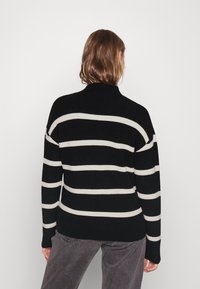 Personne aux cheveux bruns mi-longs portant un pull noir à rayures horizontales blanches et un pantalon gris, debout devant un fond clair uni.