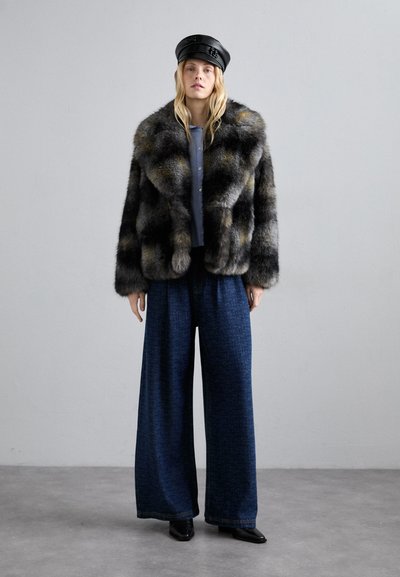 Kobieta w czarnej skórzanej czapce, szarej wzorzystej kurtce z sztucznego futra, niebieskich szerokich spodniach oraz czarnych butach na kwadratowym obcasie stoi na szarym podłożu.