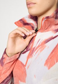 Fleece pullover met een hoge kraag, voorzien van een levendig oranje en paars abstract patroon, half-ritssluiting en een zachte textuur.