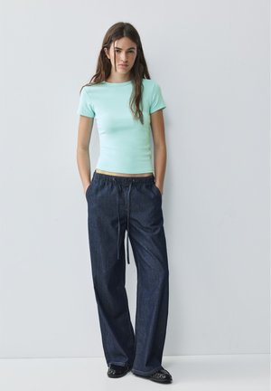 Giovane donna con lunghi capelli castani che indossa una maglietta a maniche corte azzurro chiaro e jeans larghi blu scuro con scarpe nere, in piedi con le mani nelle tasche.