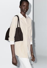 Striped button-up shirt with yellow and white lines, paired with a dark brown suede bag. Features rolled sleeves and white cuffs.