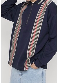 Sudadera navy con cuello de cremallera que presenta rayas verticales en crema, verde y rojo. Elaborada con un material suave y con un corte relajado.