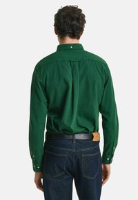 Groene katoenen blouse met lange mouwen, rugplooi-detail en knoopmanchetten. Draagt met donkere denim jeans en een zwarte riem.