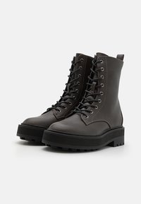 Bottines en cuir noir avec une semelle en caoutchouc plate, bout rond et plusieurs œillets pour les lacets. Elles possèdent une languette à l'arrière.