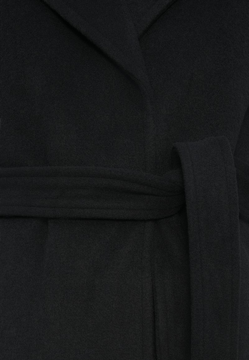 Svart fleecemantel med sjalkrage, sidofickor och matchande bälte. Den mjuka texturen och den strukturerade formen ökar värmen och komforten.
