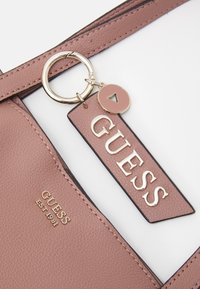 Gros plan d'un sac Guess en cuir texturé mauve avec logo doré et étiquette de porte-clés assortie accroché à la bandoulière.
