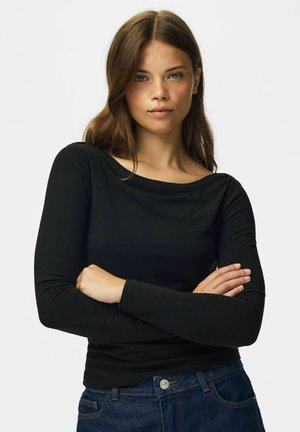 Jeune femme aux cheveux bruns, vêtue d'un haut noir à manches longues et d'un jean bleu, debout, les bras croisés, devant un fond uni.