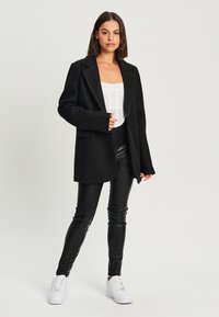 Blazer noir oversize avec des revers structurés, associé à un haut blanc et à des leggings en cuir noirs, complété par des baskets blanches.
