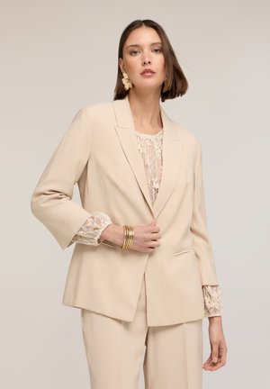 Kvinde iført beige dragt med blondebluse, guldbøjler og store blomstret øreringe, der poserer mod en ensartet neutral baggrund.