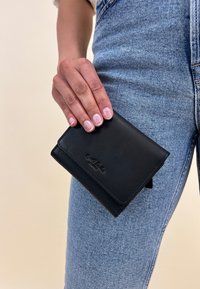 Black leather wallet held in a hand, featuring a smooth texture, rectangular shape, and embossed logo. Blue denim pants visible.